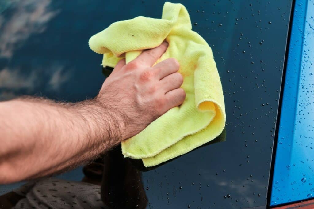 close up mans hand polishing or cleaning a rear windshield of a blue automobile with a yellow microfiber rag . manual car washing.