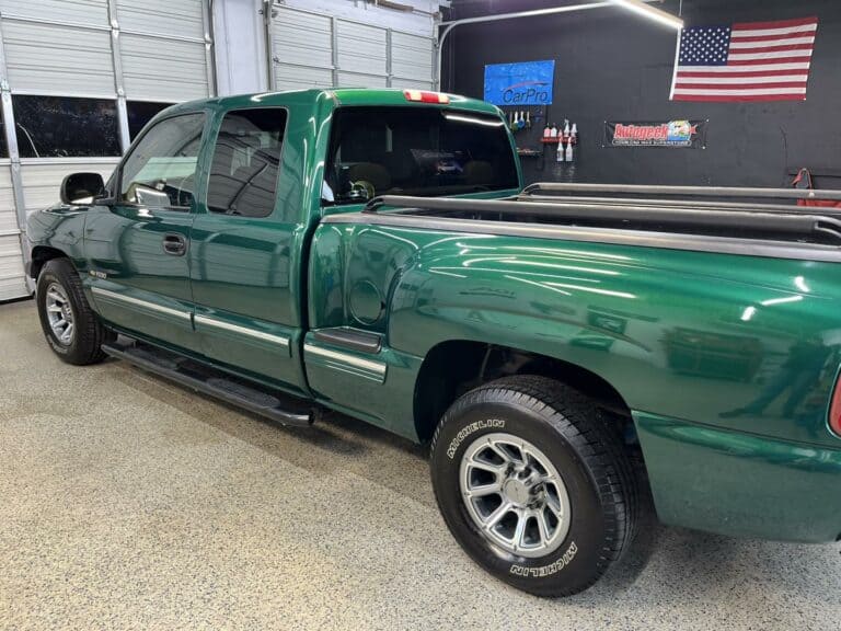 a green pickup truck is parked inside a garage with a black wall, an american flag, and various automotive signs in the background.