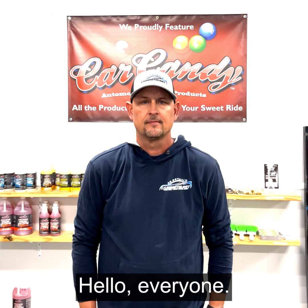 a man in a dark hoodie and cap stands in front of a "car candy" sign, with car care products on shelves behind him. the caption at the bottom reads, "hello, everyone.