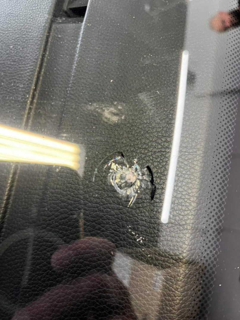 close up of a cracked windshield with a circular chip and radiating cracks, next to a blurred reflection and dashboard surface.